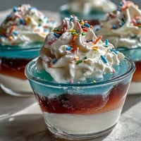 Vibrant rainbow Jello cups layered in clear cups, topped with whipped cream and colorful sprinkles for a festive dessert.